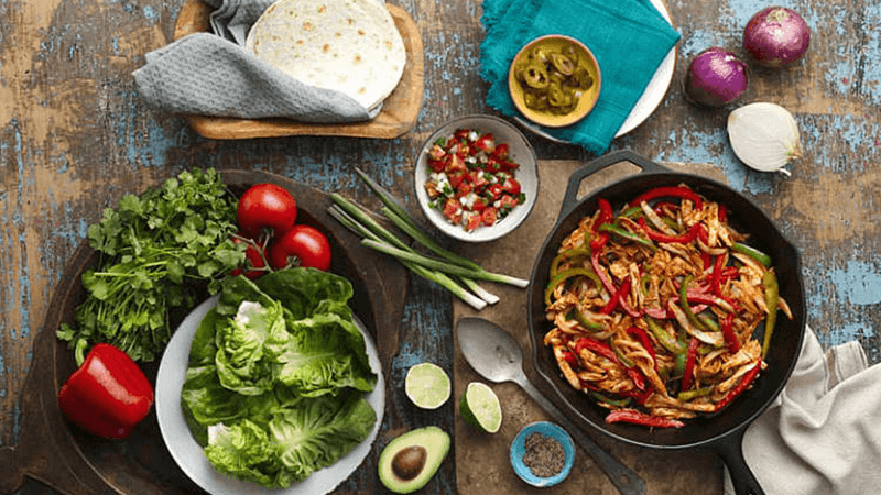 skillet filled with bbq chicken fajitas accompanied by fresh tomatoes, red pepper, halved avocado & other garnishes